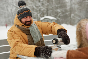 Portrait of smiling young father pouring hot tea from thermos for little girl while enjoying winter...