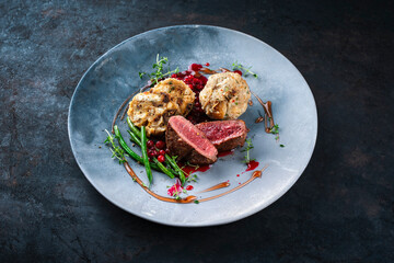 Traditionally roasted fillet of saddle of venison with bread dumplings, mushrooms and bean sticks served with lingonberry sauce as close-up on a design plate with a rustic background