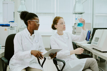 Two female scientists working on scientific research in a laboratory environment, analyzing a blue liquid solution and checking data on a tablet device