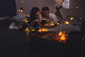 Romantic surprise for girlfriend or boyfriend. Bedroom prepared for watching old movies with popcorn, decorated with lights and candles. Cozy home Christmas atmosphere, hot chocolate with marshmallow