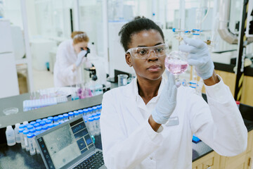 Chemist observing colored solution in modern laboratory while colleagues working in background....