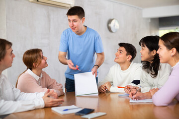 Cheerful young student discussing learning assignment with friendly coursemates gathered in...