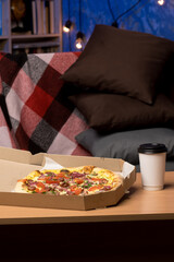 A pizza in an open cardboard box on a wooden table, topped with tomatoes, sausage, red onions, and herbs. A disposable cup with a black lid sits next to the box. Ideal for food delivery themes. 