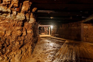 The dark and wet mine tunnel has a wooden walkway for walking through. The walls of the tunnel are made of rough-cut stone. It is lit by a single light bulb hanging from the ceiling, gold mine