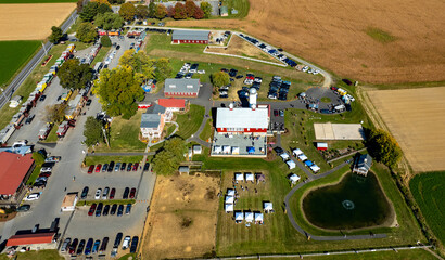 An aerial perspective showcases a bustling community event area with tents and diverse vehicles parked nearby, surrounded by lush farmland, highlighting local engagement and outdoor activities.