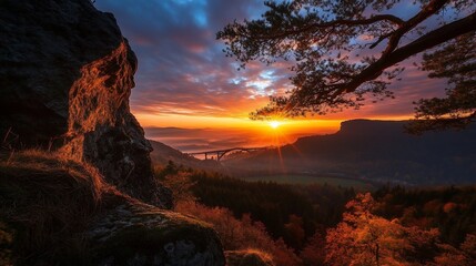 Obraz premium Sunrise over a valley with a bridge and mountains in the distance, seen through the branches of a tree.