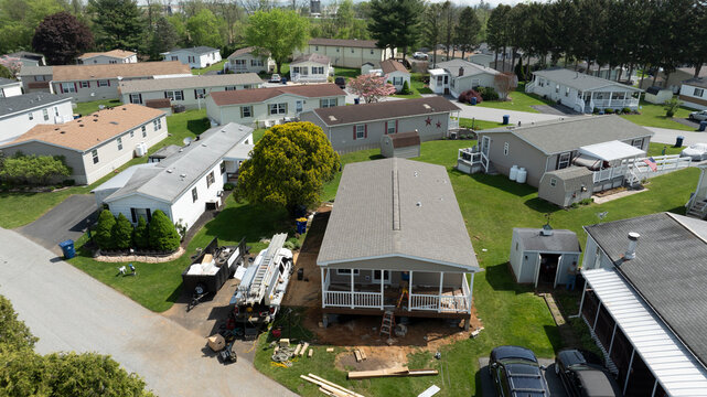 Aerial view shows mobile, manufactured homes lined along a tranquil street, surrounded by well-maintained lawns and trees. The sun casts a warm light, highlighting the neat layout and construction.