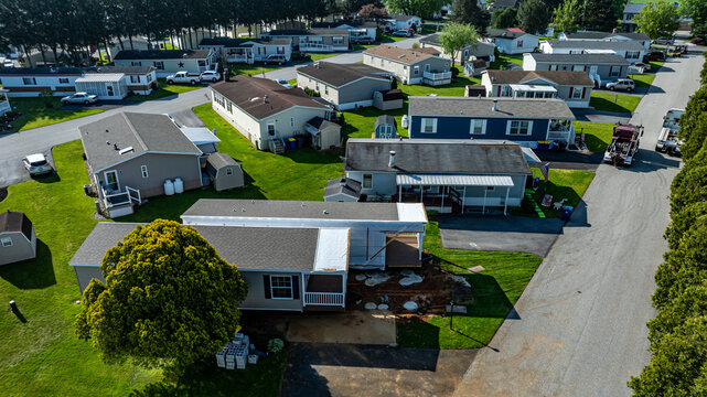 Aerial view shows mobile, manufactured homes lined along a tranquil street, surrounded by well-maintained lawns and trees. The sun casts a warm light, highlighting the neat layout and construction.