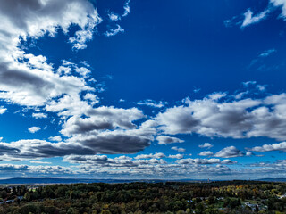 A vibrant canopy of clouds floats across a bright blue sky above a lush green forest. The autumn landscape shows hints of warm colors, creating a picturesque afternoon scene.