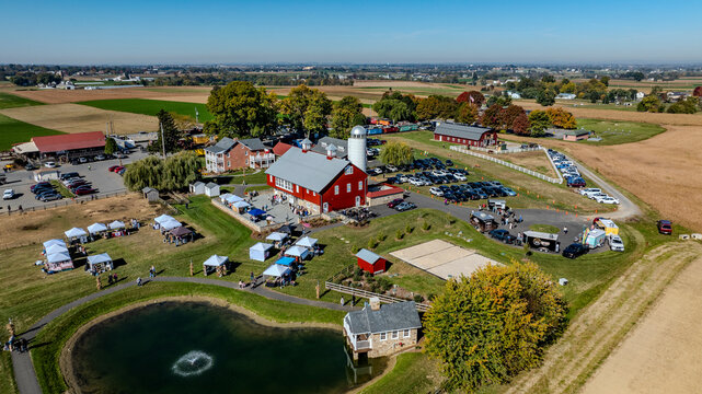 Visitors gather at a farm event featuring tents and activities around a pond, surrounded by picturesque fields and buildings under clear blue skies.