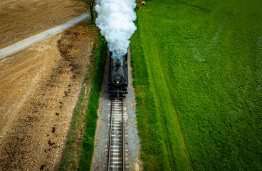 A vintage steam locomotive puffs white smoke as it moves along railway tracks surrounded by vibrant...
