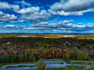 An expansive aerial view showcases a stunning autumn landscape filled with colorful trees. The vibrant foliage stretches across the countryside under a dynamic blue sky with fluffy clouds.