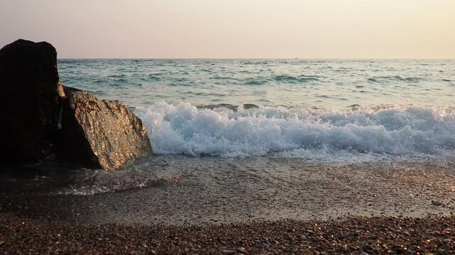 Waves gently lap at rocky shore during stunning sunset over ocean. Golden sunlight reflects off waves as they roll onto shore, creating tranquil atmosphere at sunset by sea. Montenegro summer, Canj
