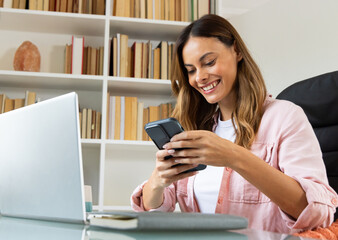 Smiling woman using smartphone at home office, working on laptop