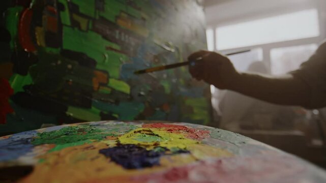 Closeup of palette with smudges of bright acrylic or oil paint, and hand of anonymous artist working on abstract masterpiece in background, in hazy light from window