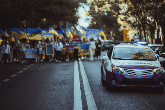 Ukrainian anti-war protest, a defocused crowd holding national flags and anti-war signs in a public square. Demonstrators showing strong resistance; selective focus on a police car on the right - Powered by Adobe