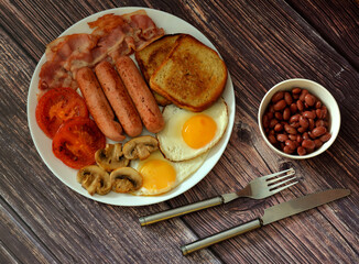 English breakfast, sausages, toast, scrambled eggs, fried bacon and mushrooms with tomatoes on a wooden table, next to cutlery and a cup of beans.