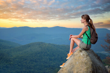 Naklejka premium Hiker girl resting on rocky mountain top enjoying morning nature during her travel on wilderness trail. Lonely female traveler traversing high hilltop route. Healthy lifestyle concept