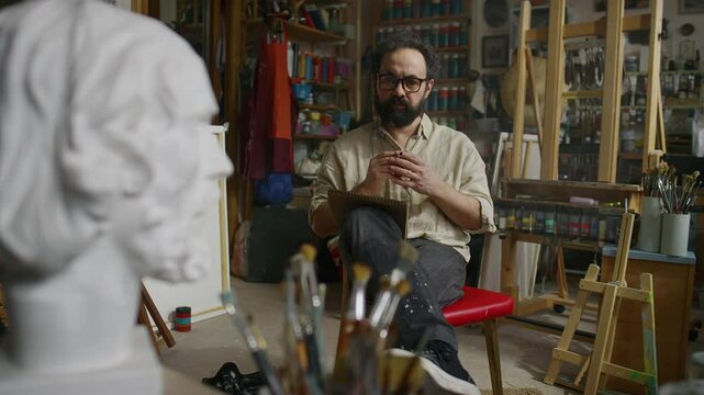 Full shot of mature curly-haired, bearded Caucasian male painter in glasses sitting on chair in studio, studying white alabaster head sculpture, drawing sketch in notepad, while smoking cigarette