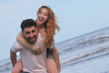 young cheerful couple goofing around having fun with piggyback ride on the beach  on summer day