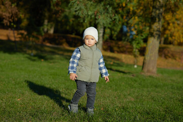 A young child stands on green grass in an autumn park, dressed warmly in a beanie, plaid shirt, and vest, looking curiously at the camera. The background is filled with fall foliage.