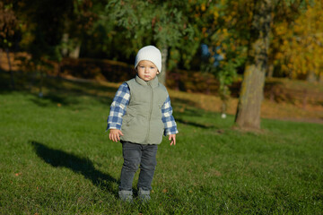 A young child stands on green grass in an autumn park, dressed warmly in a beanie, plaid shirt, and vest, looking curiously at the camera. The background is filled with fall foliage.