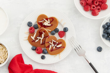 A heap of heart shaped pancakes with berries and a fork on the white plate on grey background