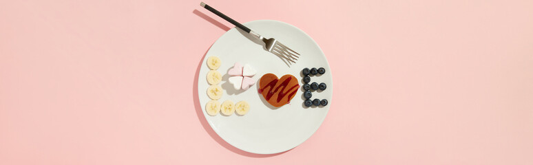 A Love symbol made of heart shaped pancake, marshmallow and fruits on the white plate with fork