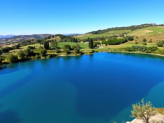 Fototapeta premium Clear blue lake nestled in the tranquil countryside of Rome under the bright midday sun, surrounded by lush greenery and distant rolling hills, countryside, scenic