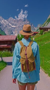 An attractive girl in a blue shirt and a fashionable hat spends her holidays alone, traveling through the Alpine meadows. Tourist with a backpack travels through Switzerland with incredible views