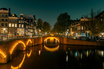 Amsterdam’s historic canal bridges illuminated at night. The Keizersgracht and Reguliersgracht intersection glows romantically, reflecting typical Dutch architecture and city lights in the water.