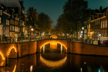 Amsterdam’s historic canal bridges illuminated at night. The Keizersgracht and Reguliersgracht intersection glows romantically, reflecting typical Dutch architecture and city lights in the water.