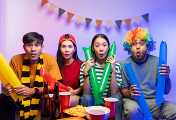 Group of asian friends man and woman watching cheering football match on TV at home. Eating snack and drinking beer. Sport fans people shouting and celebrating sport team victory match.