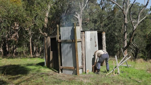 Australian pioneer bringing firewood into his tin humpy with smoke blowing out the chimney.