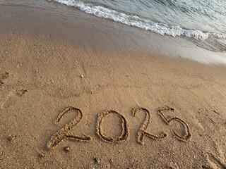 Capturing the coming year '2025' handwritten in the beach sand, with clear imprints visible on the golden surface, accompanied by gentle waves lapping at the shore.