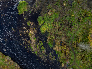 Top-down aerial photo of the Jagala River near Jägala Waterfall in Estonia. On the left the river flows with dark, almost black water speckled with white foam. On the right rugged green landscape.