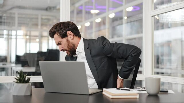Tired businessman sits at his desk and holds his lower back in pain. He's experiencing discomfort after sitting for too long. Office work hazards, back pain, and sedentary lifestyle concept.
