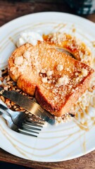 A plate of French toast topped with crumbs and a drizzle of caramel sauce, accompanied by a dollop of whipped cream and a side of nuts. A fork and knife rest beside it on the plate.

