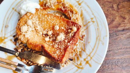A plate of French toast topped with crumbs and a drizzle of caramel sauce, accompanied by a dollop of whipped cream and a side of nuts. A fork and knife rest beside it on the plate.
