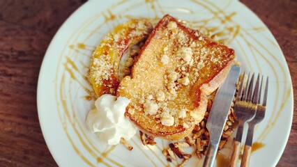 A plate of French toast topped with crumbs and a drizzle of caramel sauce, accompanied by a dollop of whipped cream and a side of nuts. A fork and knife rest beside it on the plate.
