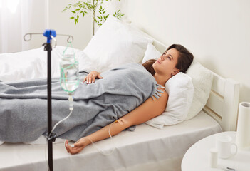 Young upset woman lying on bed at home or in medical clinic while receiving IV drip infusion and...