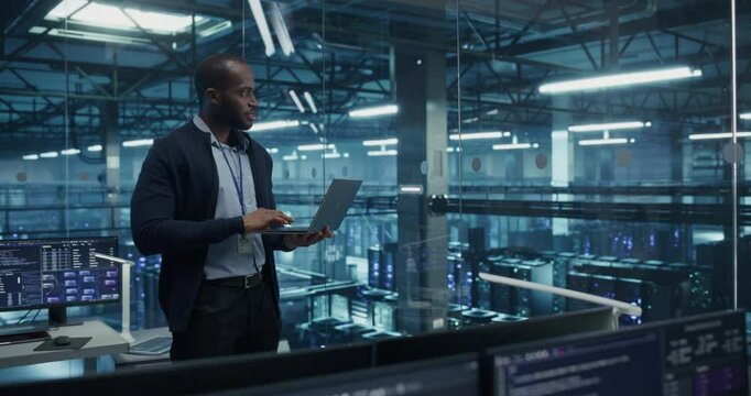 Black Male Software Developer Analyze System Performance Metrics on a Laptop Inside a Modern Data Center. Young Manager Ensuring Efficient Operations for Critical Infrastructure