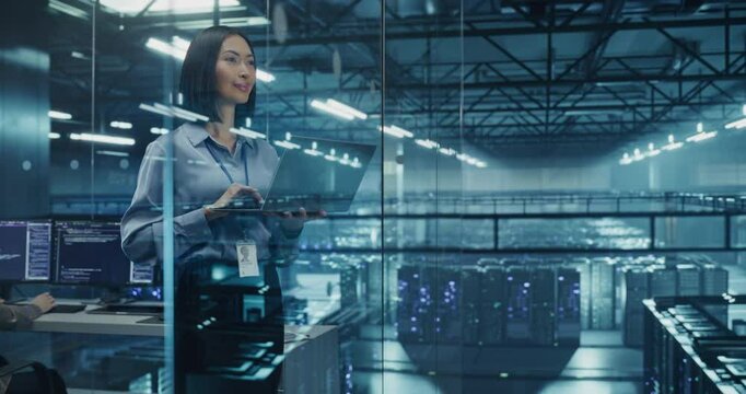 Asian Female Data Analyst Studies Traffic Logs on Her Laptop Computer, Standing in a Modern Data Center with Server Systems. Young Woman Searching for Patterns to Improve Online Data Flow