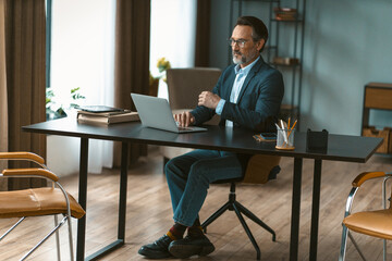 Man is sitting at a desk with a laptop and a pen. He is wearing a suit