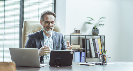 Man is sitting at a desk with a laptop and a tablet. He is wearing a suit and glasses