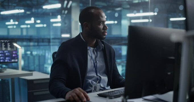 Portrait of a Dedicated Data Scientist in a Data Center Office, a Black Man Concentrates on His Work on a Computer, Managing System Security and Processing Key Information Online