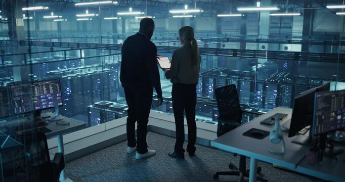 Male DevOps Engineer and Female IT Consultant Work on a Tablet Computer, Standing in Office Above the Rows of Servers in a Data Center, Working on Research and Development of Artificial Intelligence
