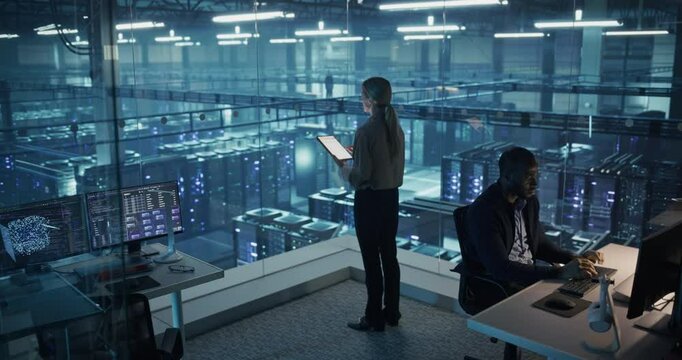 Female Cybersecurity Expert Standing with Her Back to Camera, Works on Her Tablet in a Modern Facility, Running Diagnostic Tests to Identify Network Vulnerabilities in a Data Center with Server Racks