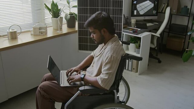High angle view of young Indian engineer in wheelchair working on laptop in modern sustainable office