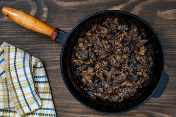 Cast iron skillet with fried wild mushrooms with onions and sour cream on wooden background, top view, closeup